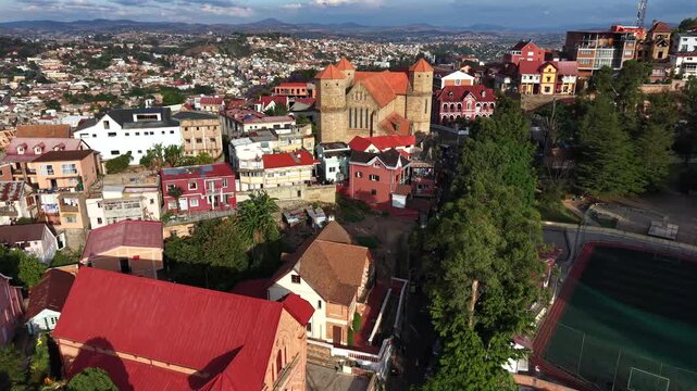 Aerial view of colorful hillside town with historic church and winding streets. Red roofs contrast with green trees and distant mountains. Quiet atmosphere, no visible people