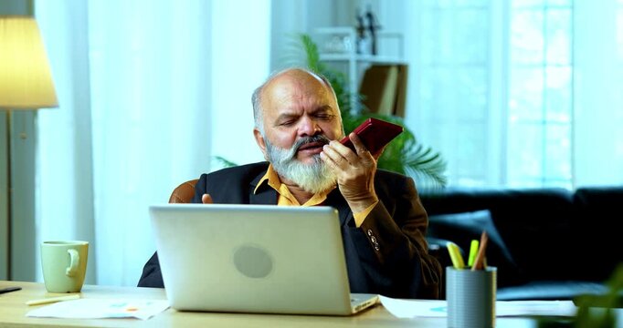 Indian senior businessman or corporate executive multitasking by speaking on speakerphone via his smartphone while actively typing on a laptop at his desk in a cozy office with moody lighting.