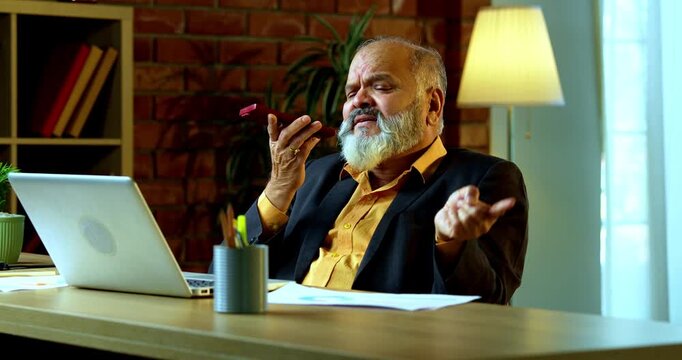 Indian senior businessman or corporate executive multitasking by speaking on speakerphone via his smartphone while actively typing on a laptop at his desk in a cozy office with moody lighting.