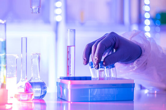Gloved hand holding test tube in modern laboratory with sample vials and glassware under blue light, concept of research, biotech and clinical testing.