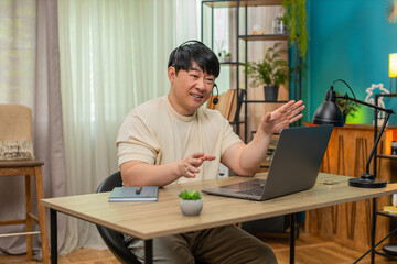 Asian man sitting at home table speaks on laptop headset video call, providing customer support in...