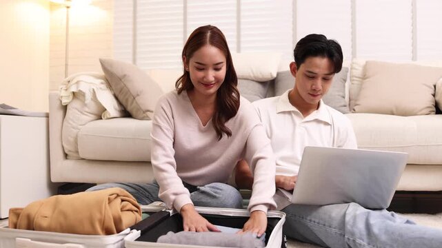 Young Asian couple prepares for a trip at home, The woman folds clothes into a suitcase while the man uses a laptop to book travel or check flight details in their cozy living room