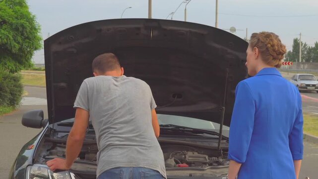 Young woman in blue suit stands besides man and help him. He is at opened front body of car. Guy try to repair car. He takes one wrench after another. Young woman gives it to him.