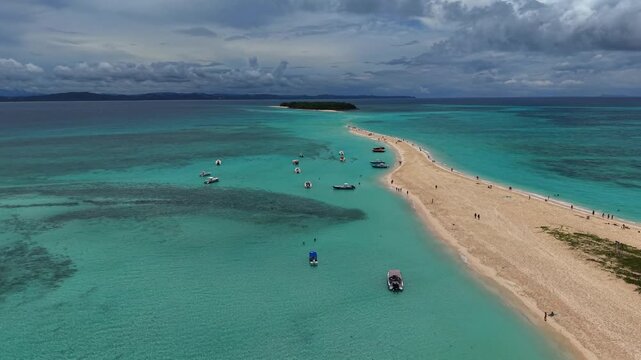 Aerial view of a long, sandy beach curving into turquoise waters. Several boats float near shore, people stroll along the sand. Sky is cloudy, hinting at an approaching storm