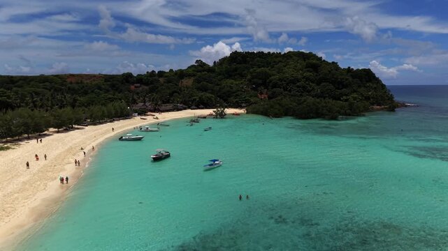 Aerial view of tropical beach with turquoise waters. White sand curves around lush green hills. Small boats float near shore. People relax, swim, and sunbathe peacefully. Ideal for vacation, travel