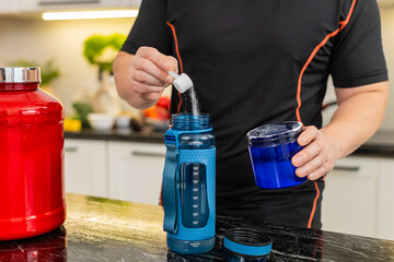Naklejka premium Man adds supplement powder into shaker bottle in kitchen preparing isotonic or creatine drink. Shaking bottle mixes powder with water for workout hydration muscle recovery energy fitness routine gym.