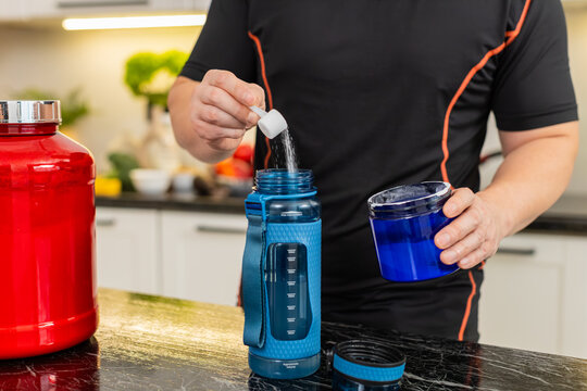 Man adds supplement powder into shaker bottle in kitchen preparing isotonic or creatine drink. Shaking bottle mixes powder with water for workout hydration muscle recovery energy fitness routine gym.