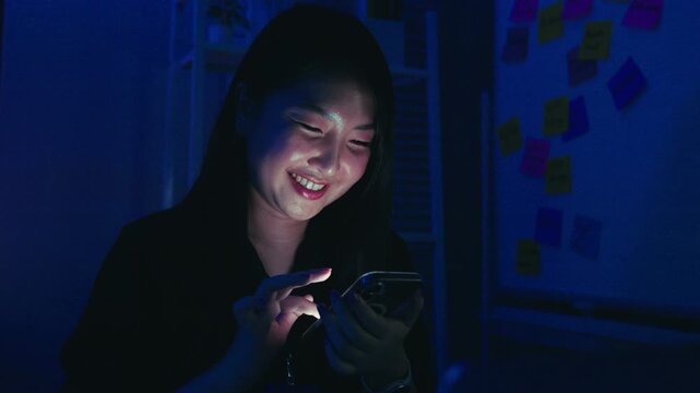 Young Asian woman using smartphone at night with screen light illuminating her face in dark room