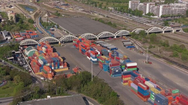Shipping Container Port Aerial View: An extensive shipping container port, viewed from above, showcases rows of stacked cargo containers under arched bridges and nearby railway lines.