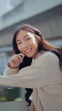 Happy Asian Gen Z woman smiling outdoors in the city. Beautiful young female with long hair enjoying a sunny day, showcasing wellness, positive emotions, and a modern lifestyle.