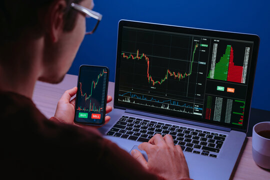 Over shoulder view of crypto trader analyzing live crypto and stock charts with trading platform app, displayed on his laptop and smartphone screen in dark neon lit room.
