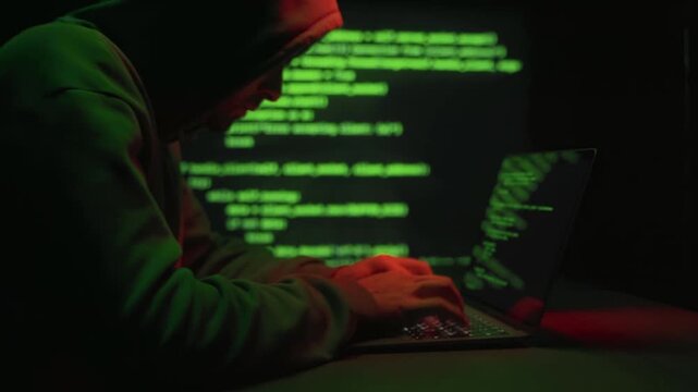Anonymous hacker in hoodie working on computer with glowing green code background. Hacking process, cybercrime, phishing, password theft and digital intrusion.