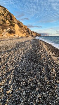 Pebble beach, sunset, ocean waves as a solitary traveler walks along the beautiful coastline with dramatic cliffs and tranquil sky. Vertical video