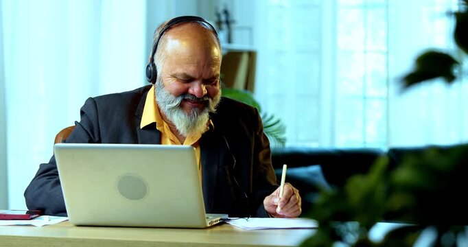 Indian senior businessman or executive wearing a headset with a microphone, taking handwritten notes while actively working on his laptop at a desk in a cozy office with professional moody lighting.