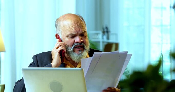 Indian senior businessman or corporate executive multitasking by reviewing paper documents while speaking on a smartphone and working on a laptop at his desk in a cozy office with moody lighting.