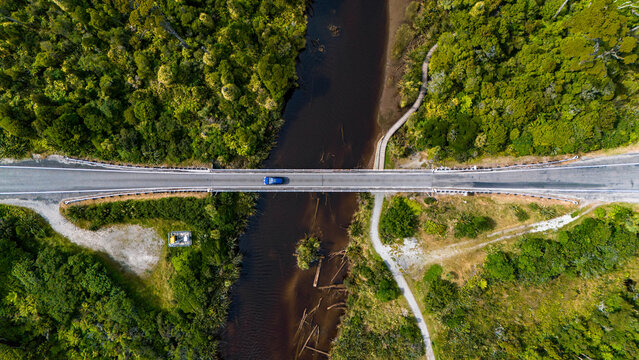 Aerial view of a single blue car crossing a bridge over the dark, winding river, framed by vibrant green forest, Ship Creek, Haast, West Coast Region, New Zealand.