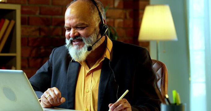 Smiling Indian senior businessman or corporate executive wearing a headset with a microphone, taking handwritten notes while working on his laptop at a desk in a cozy office with moody lighting