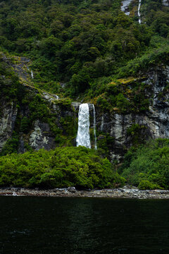 View of a powerful waterfall cascading down the rugged, verdant cliffs into the dark, reflective waters, creating a serene yet dramatic landscape, Milford Sound, Southland Region, New Zealand.