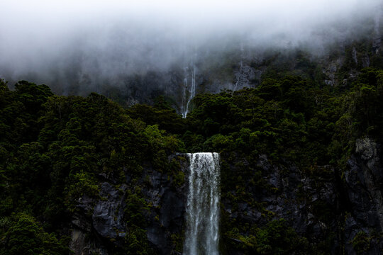View of a dramatic waterfall cascading down a dark, rocky cliff face shrouded in mist and lush vegetation, creating an ethereal atmosphere, Milford Sound, New Zealand.