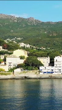 Picturesque coastal village with traditional buildings nestled between the sea and lush green mountains in Erbalunga, Corsica, on a sunny day.