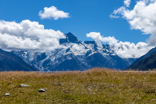 View of snow-capped mountains piercing the bright blue sky, partially veiled by puffy white clouds, above a field of golden grass, Mount Cook, Canterbury Region, New Zealand.