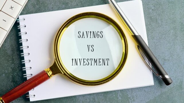 A golden magnifying glass reveals the words savings versus investment on a white notebook on a grey desk