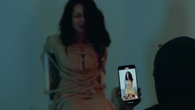 Male kidnapper filming a video with a hostage on his phone. Woman sitting on a chair and screaming, tied with a rope. Kidnaping concept. Slow motion