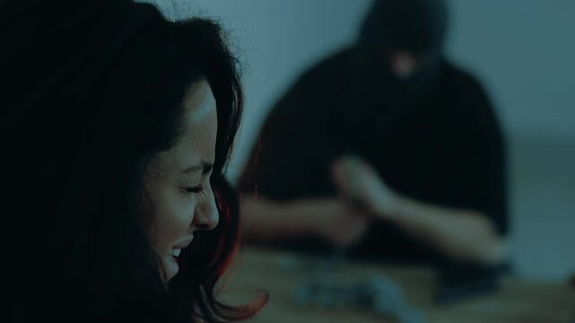 Scared young woman crying sitting in the basement, the kidnapper in a black balaclava in the background. Kidnaping, crime, violence concept. Slow motion