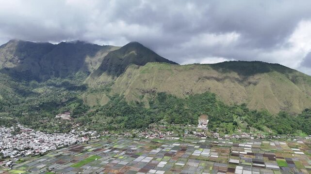 Aerial view of a stunning mountain landscape with lush green peaks, a tranquil village, and a vast expanse of patchwork agricultural fields under a dramatic cloudy sky.