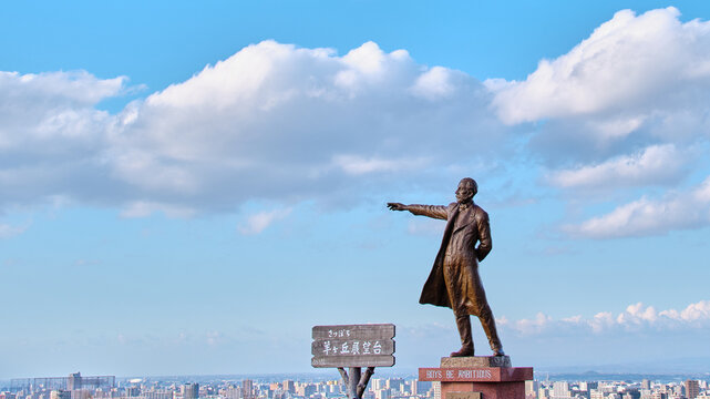 春の青空と雲を背景にした羊ヶ丘展望台のクラーク博士像と札幌の街並み  Statue of Dr. Clark and Sapporo Cityscape at Hitsujigaoka Observation Hill Against a Spring Blue Sky