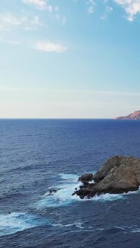 Waves crash against a rugged rocky coastline under a clear blue sky near Cargese, Corsica. Tranquil seascape with vibrant ocean colors and dramatic cliffs.
