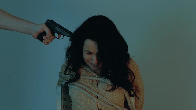 Male kidnapper put a gun to the temple of the hostage. Woman sitting on a chair and tied with a rope. Crime, violence, robbery and killer concept. Slow motion