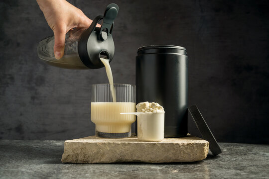 Pouring protein vanilla shake in drinking glass and scoop with whey protein powder on dark background