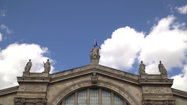 Low angle of the Gare du Nord facade with ornate statues and arch window against a blue sky with white clouds.