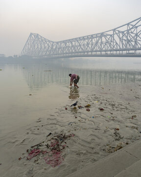 View of a man wading through the muddy riverbank under the majestic Howrah Bridge, casting a long shadow on the water in Kolkata, West Bengal, India.
