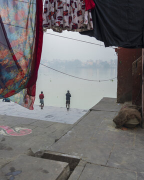 View of two figures standing on a concrete platform by the misty river, framed by colorful fabrics drying overhead in Kolkata, West Bengal, India.