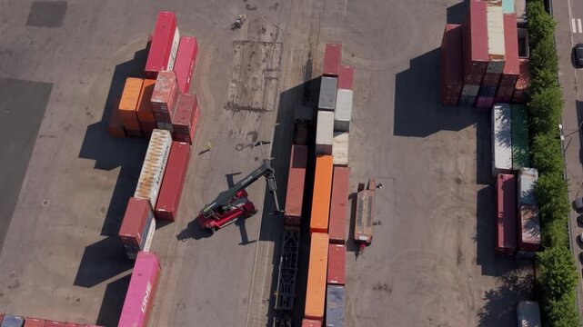 Drone aerial of an industrial freight terminal in Italy. Reach stacker and terminal tractor vehicles actively handling and transporting shipping containers within a logistics yard.