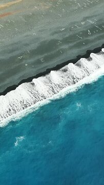 Aerial shot of turquoise waves crashing on the dark sandy shore of Plage de Nonza, Corsica. The unique coastline features white sea foam and clear blue water.