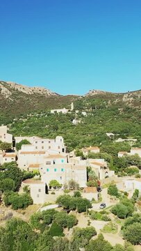 Scenic aerial shot of the hilltop village of Pigna surrounded by lush greenery and mountains under a clear blue sky in Corsica, France.