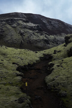 View of a dark path winding through mossy green hills under a cloudy sky, leading towards a rugged mountain in Skaftarhreppur, Iceland.