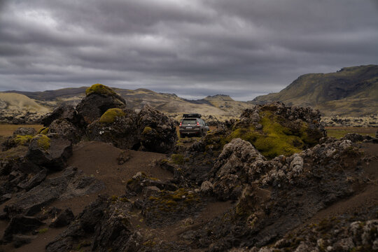 View of a rugged landscape with moss covered rocks under a cloudy sky with a vehicle in the distance, Skaftarhreppur, Iceland.