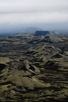 View of a mossy lava field stretches under a cloudy sky, with distant mountains fading into the horizon, in Skaftarhreppur, Iceland.