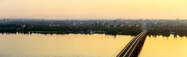Obraz premium Dnipro, Ukraine cityscape during golden hour, an aerial panorama showcasing the Dnieper River and a major bridge bustling with transport vehicles, reflecting warm sunlight across the tranquil water su