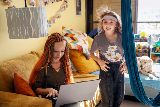Mother working on a laptop while her active daughter jumps nearby in a living room.  Working mother