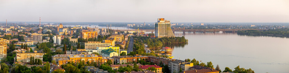 Obraz premium Dnipro, Ukraine, panoramic cityscape at sunrise, showcasing the majestic Dnieper River flowing through, highlighted by several bridges, unique modern and residential architecture, lush green spaces, a