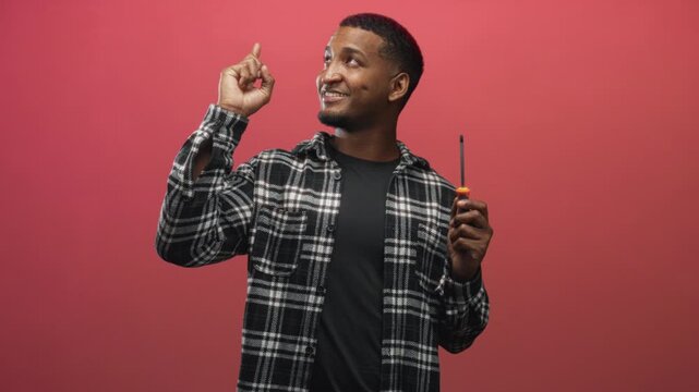 Young black man holding a screwdriver and pointing finger up in a studio with red backdrop and plaid shirt; optimism repair.