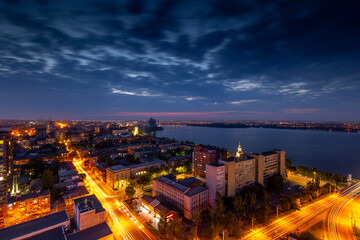 Fototapeta premium Dnipro, Ukraine, aerial twilight view showcases a bustling cityscape along the Dnieper river, illuminated by vibrant streetlights and car light trails, under a dramatic cloudy sky Includes copy space