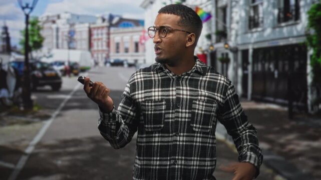Man holding car keys and keyfob with hand raised in an open questioning gesture on a street near a storefront and building with flag, wearing glasses and plaid shirt; confusion.