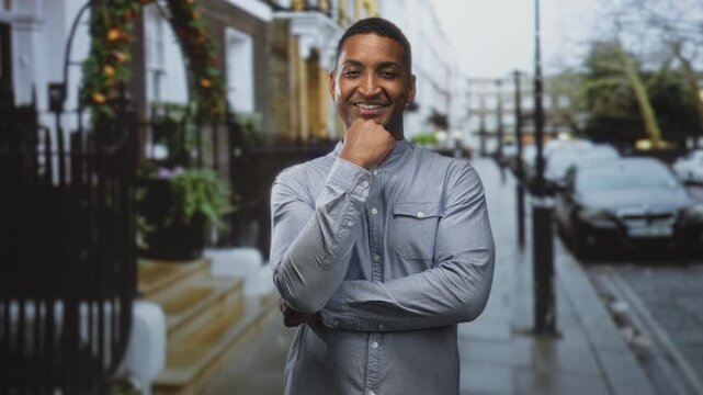 Man with hand on chin smiling, crossed arms and casual shirt visible on a street with town steps and parked cars; confidence success.