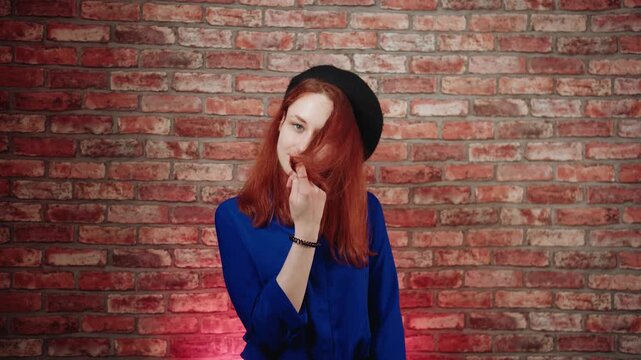 A portrait of a playful young caucasian redhead woman in a blue blouse and black beret, flirts and playfully twirls a lock of hair against a brick wall. A gesture of sympathy, interest and curiosity